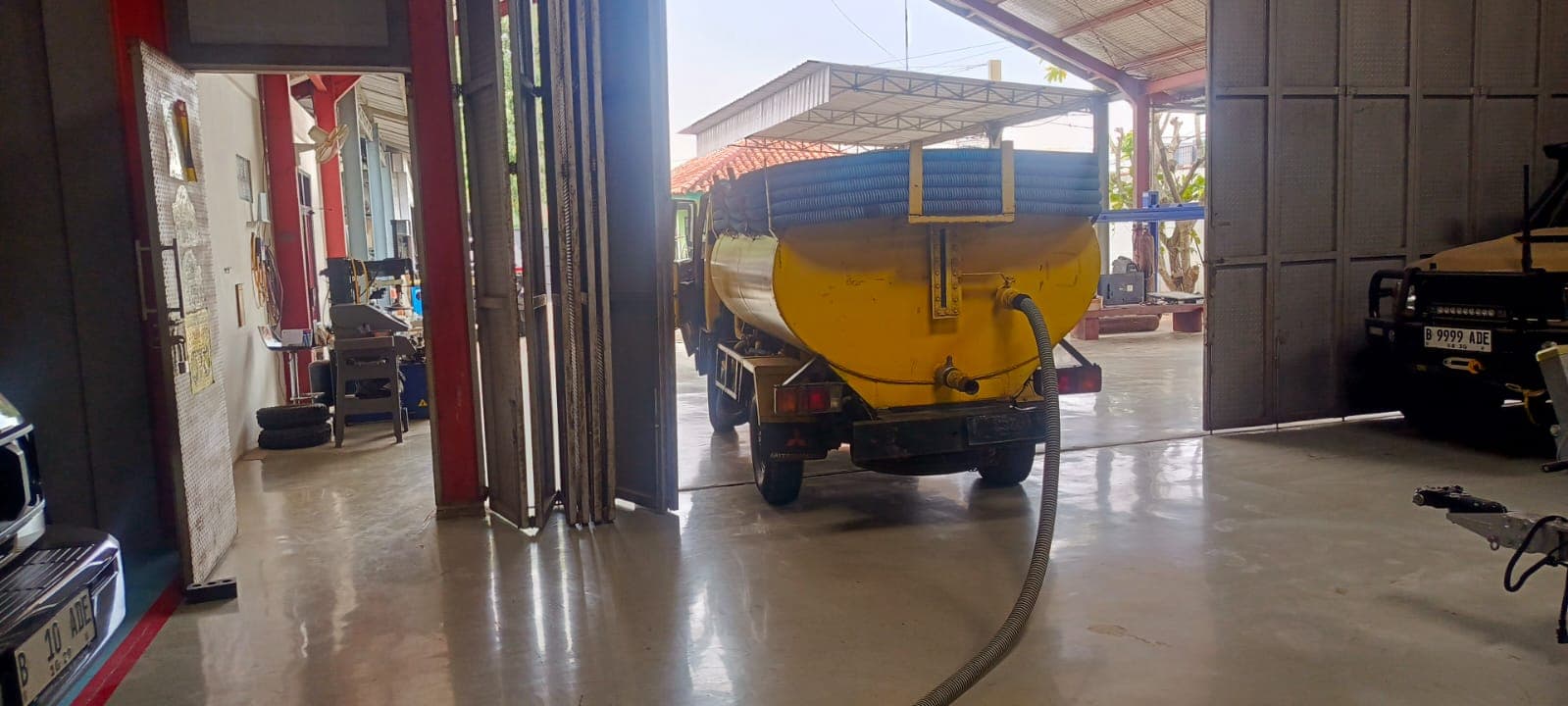 Truk tangki kuning dengan selang panjang terparkir di dalam bengkel dengan pintu terbuka lebar.